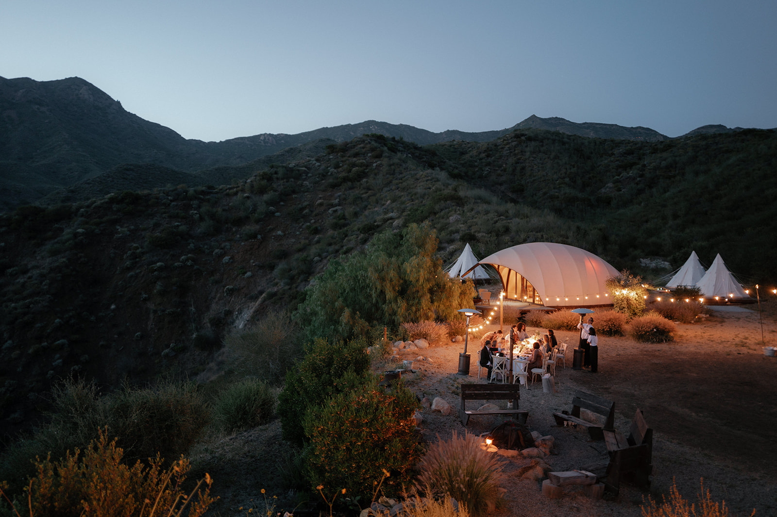 Wild Malibu Film Retreat wedding reception at dusk with string lights, mountain landscape, and guests gathered outdoors in Malibu